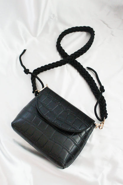 A black rectangular crossbody flap bag with pleated straps. 